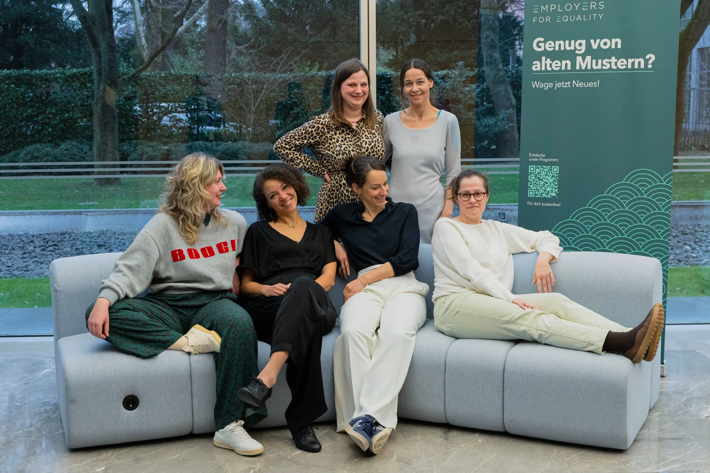 Fünf Teilnehmerinnen sitzen und stehen lachend um ein Sofa vor einem Employers-for-Equality-Rollup.